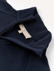 A navy blue Merino Kids AirWool Half Zip shirt is shown partially unzipped, revealing a beige tag with red stitching and text inside the collar. The AirWool 260 fabric appears soft and slightly textured.
