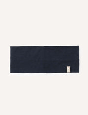 A flat, rectangular navy merino wool headband, featuring a small beige tag on one edge, is displayed against a plain white background. The Headband – Merino Kids Light offers versatile comfort for everyday use.