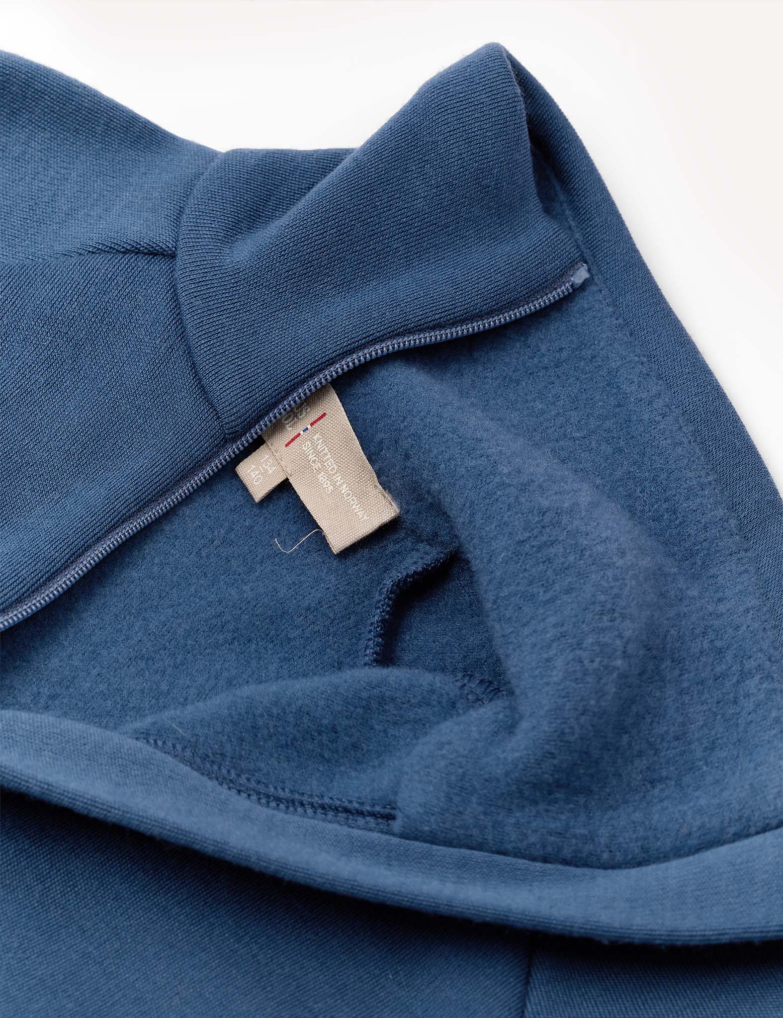 Close-up of the Jacket – Merino Kids AirWool in blue, crafted from brushed wool. The image highlights its soft inner lining and a beige fabric tag adorned with small red and white accents. The jacket is partially unzipped to reveal the cozy interior material.