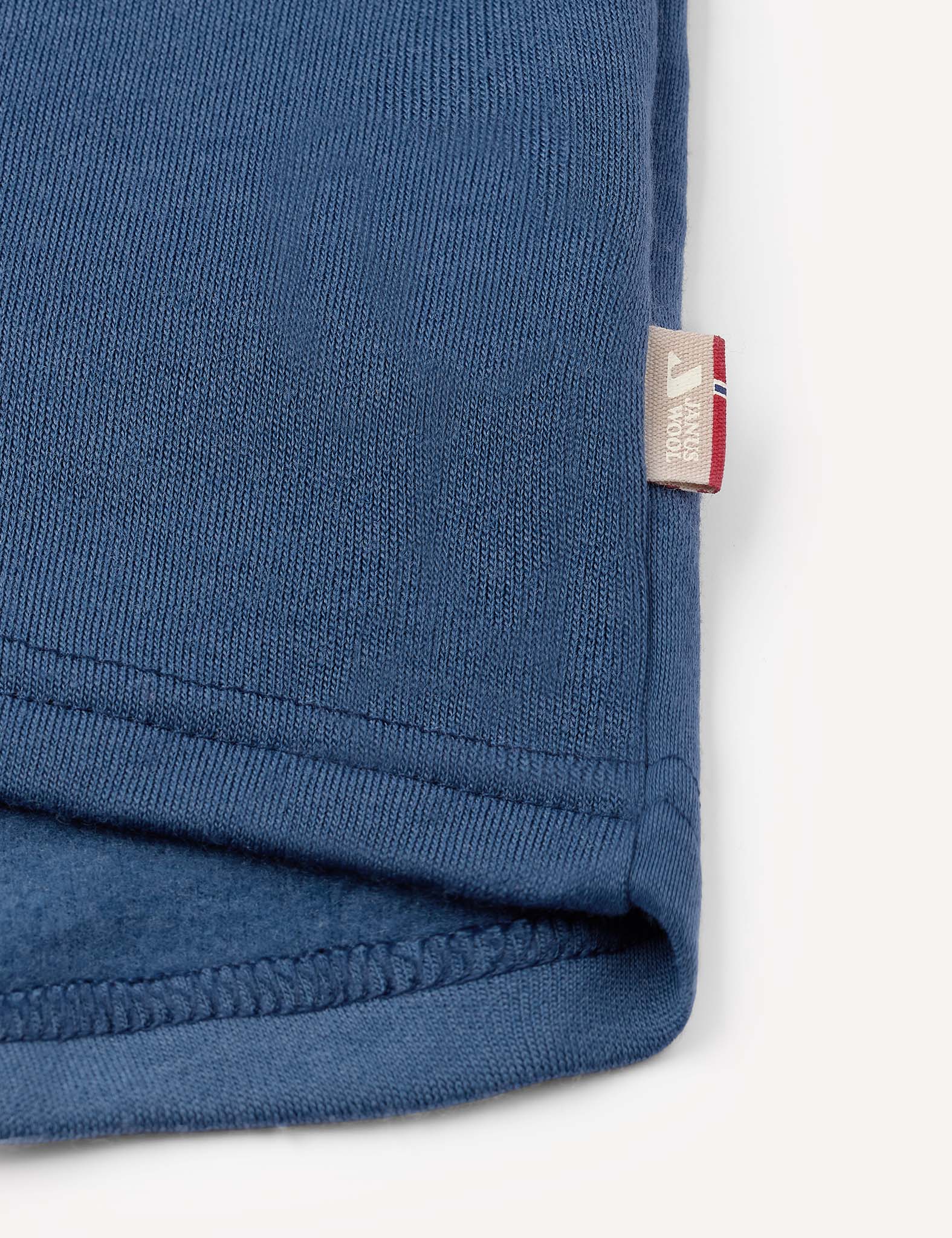Close-up of the hem of the Jacket – Merino Kids AirWool in blue, featuring a small beige tag with text and a red, white, and blue accent on the edge, set against a plain background.