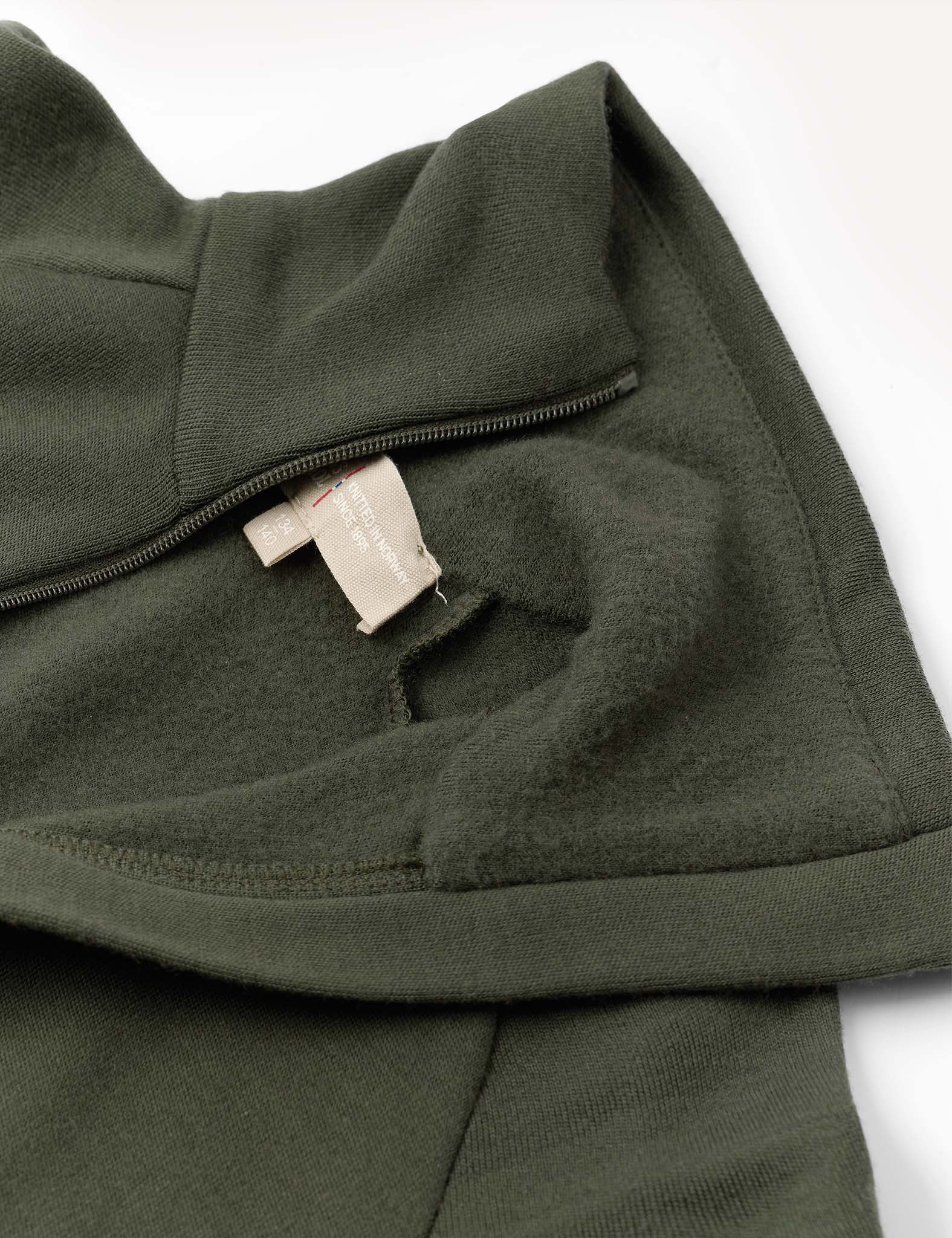 Close-up of the olive green Jacket – Merino Kids AirWool, featuring a flexible wool design with a partially unzipped collar, soft inner lining, and two beige fabric tags sewn into the neckline. The photo highlights the garment’s brushed AirWool texture and detailed construction.