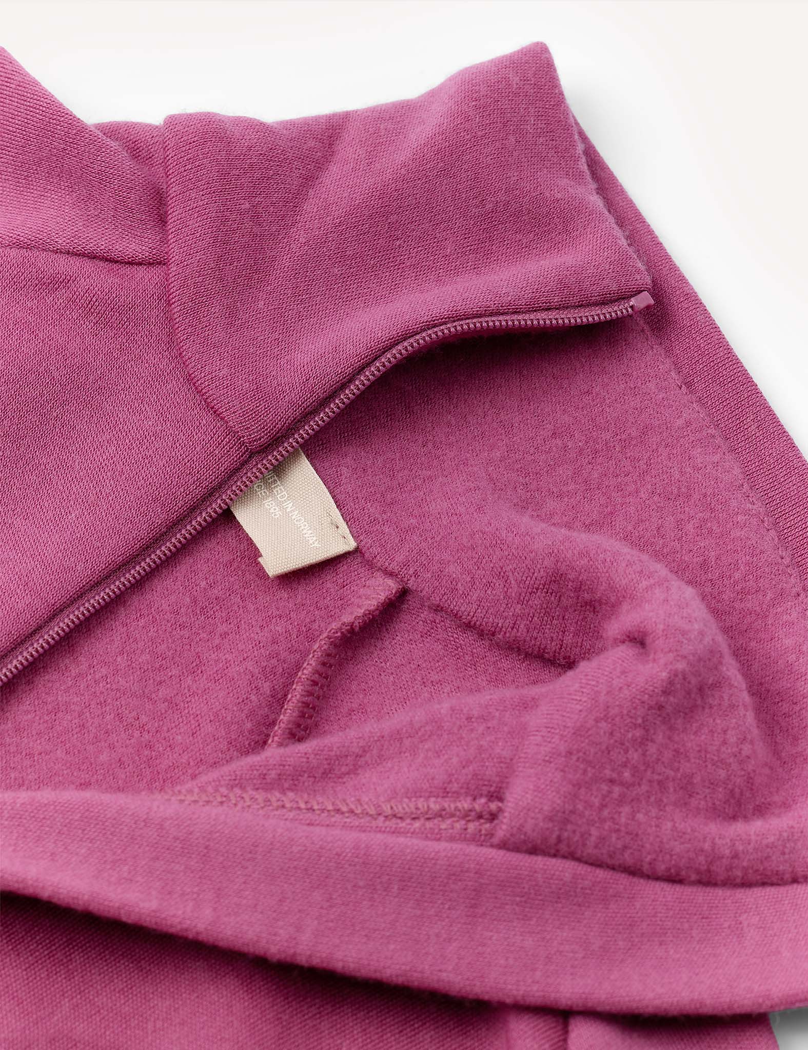A close-up of the Jacket – Merino Kids AirWool in a mauve or pinkish-purple shade highlights its soft brushed AirWool fabric texture, with an off-white clothing tag partially visible near the seam.