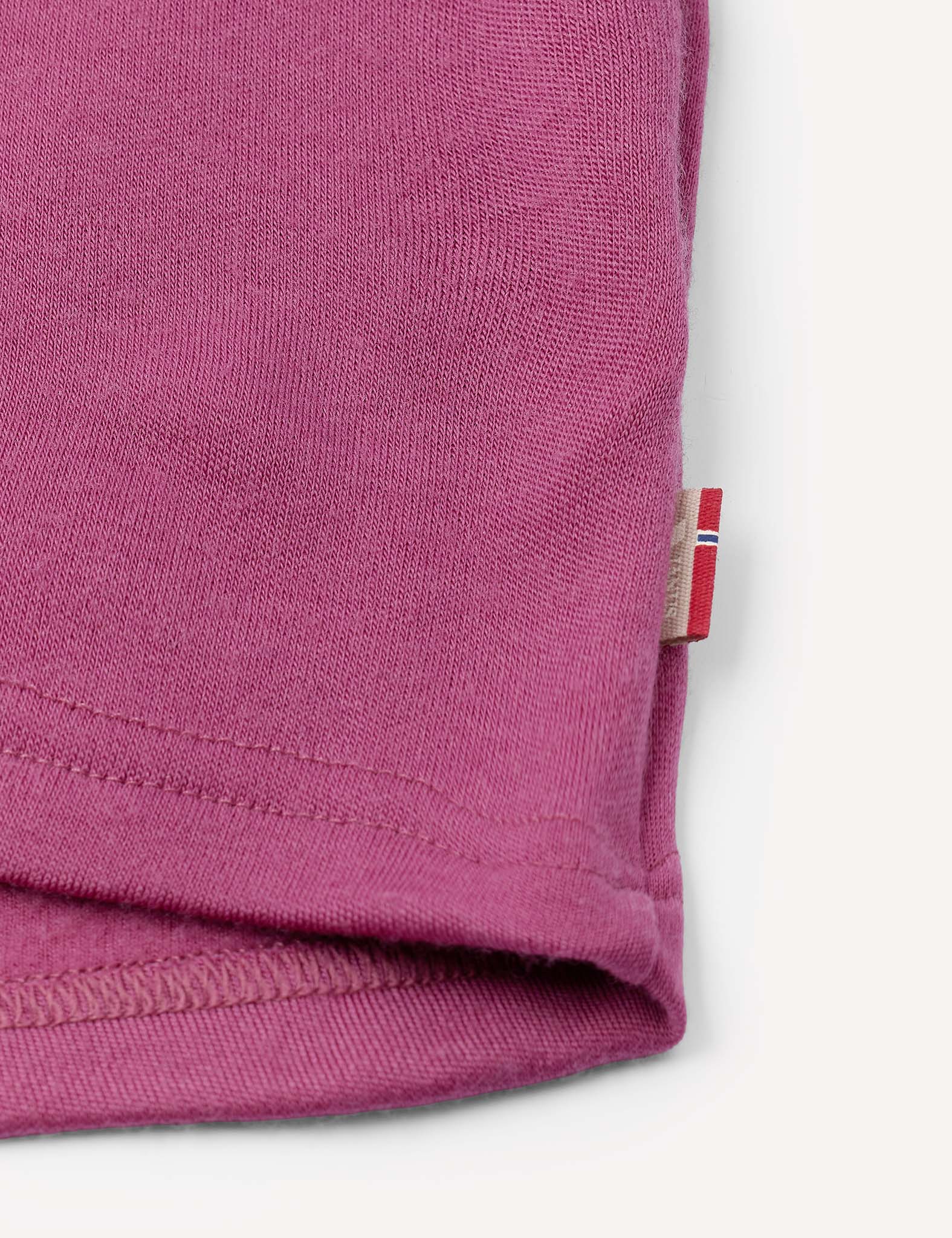 Close-up of a hemmed edge of a pink fabric, likely a t-shirt sleeve, featuring a small tag with a red and white stripe pattern sewn into the seam, highlighting details reminiscent of the Jacket – Merino Kids AirWool craftsmanship.
