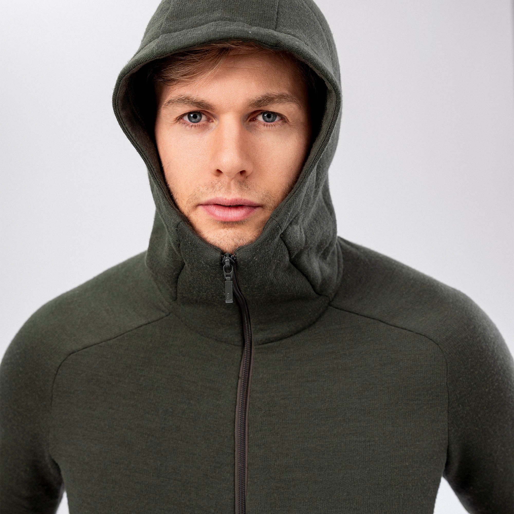 A man wears the Hooded Jacket Merino – AirWool 280 Men with the dark green hood up, looking directly at the camera against a plain light gray background.