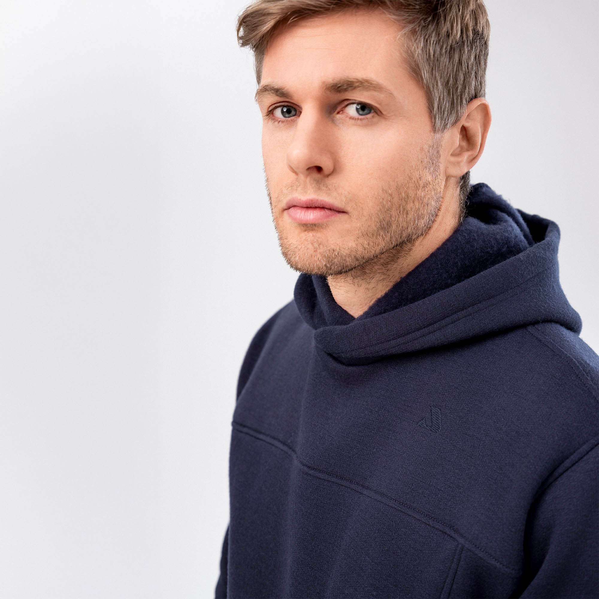 A man with short, light brown hair and stubble wears the Hoodie Merino – AirWool 280 Men in navy blue, looking slightly past the camera against a plain, light grey background.