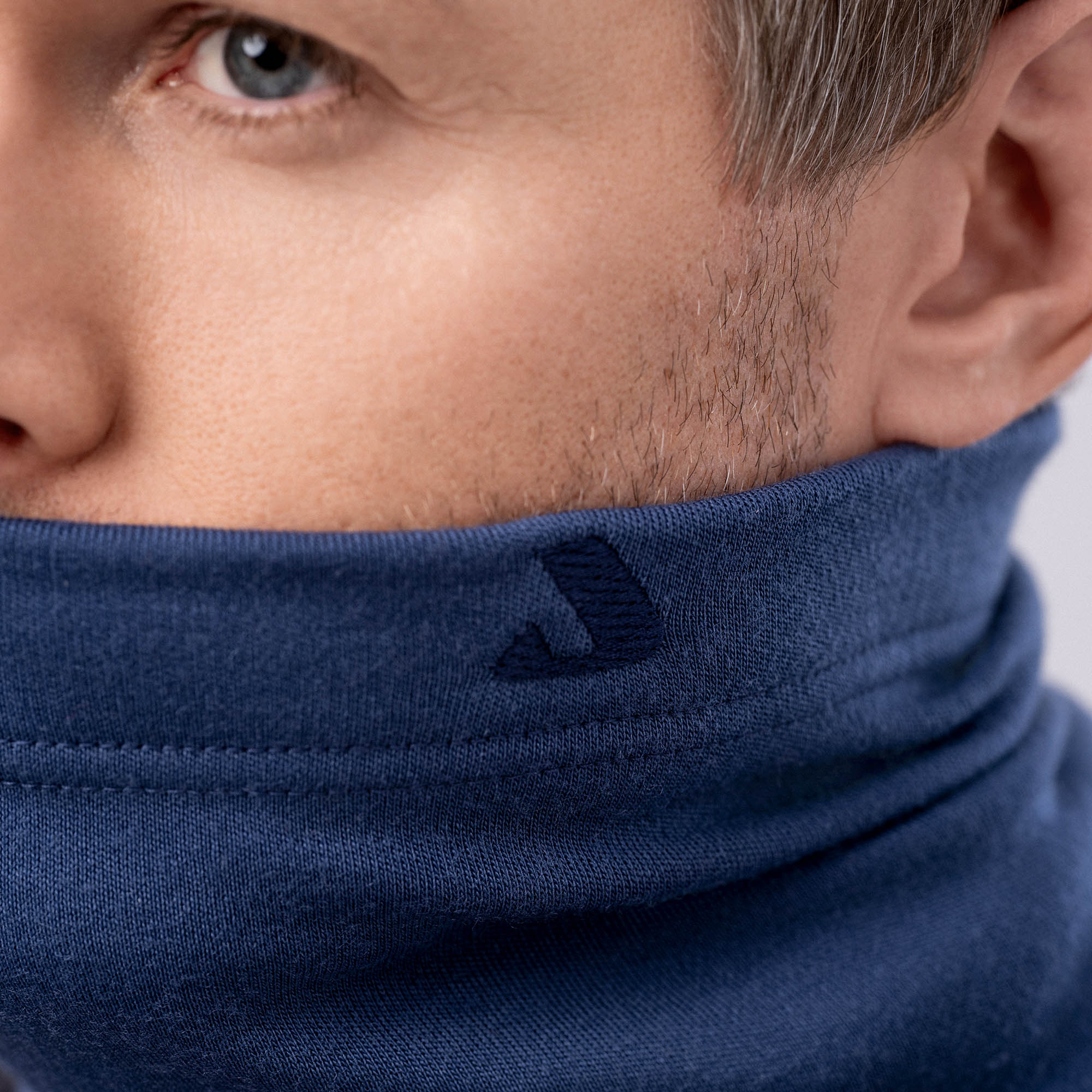 Close-up of a man wearing the Neck Warmer Merino – AirWool 260 in blue, covering his mouth and chin for warmth and breathability. The fabric displays a small, dark embroidered "J" by the collar; background is softly blurred.