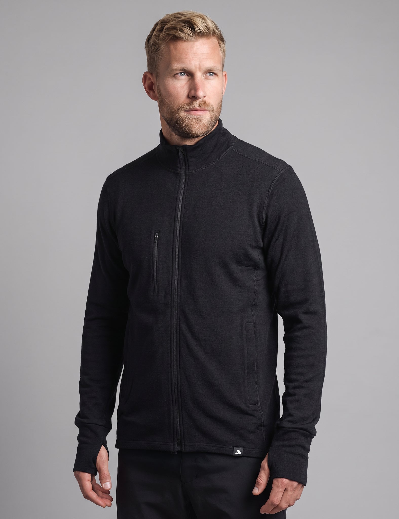 A man with blond hair and a beard wears the Norse Merino Unisex Double Layer Jacket in black along with black pants, standing against a plain gray background and looking slightly to the side with a neutral expression.