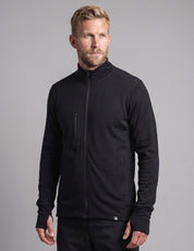A man with blond hair and a beard wears the Norse Merino Unisex Double Layer Jacket in black along with black pants, standing against a plain gray background and looking slightly to the side with a neutral expression.