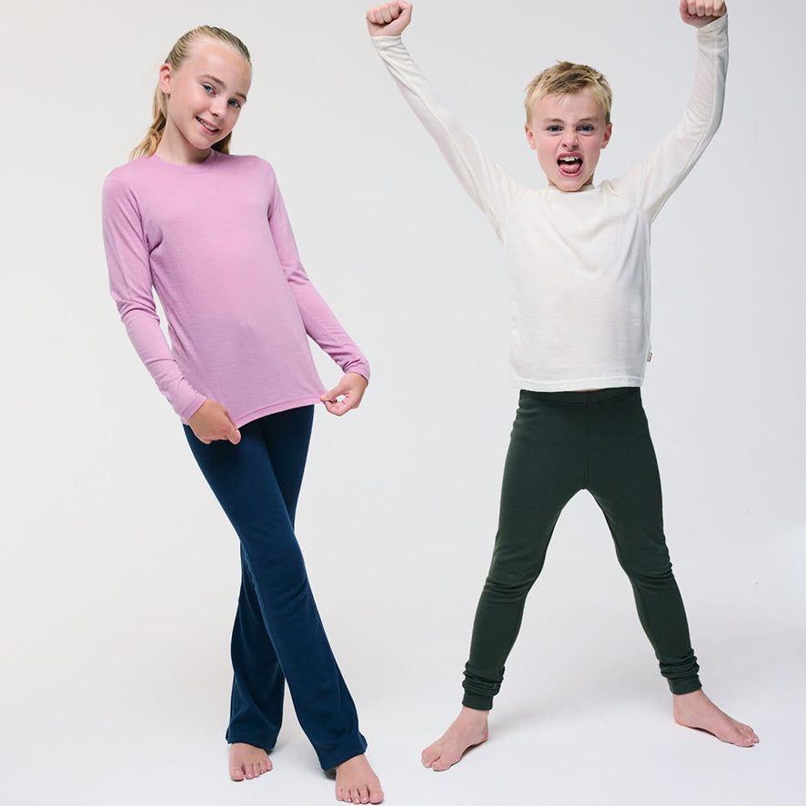 A girl in a pink merino wool top and blue pants stands smiling, while a boy in a white top and green pants stands barefoot with arms raised and a playful expression. The background is plain white.