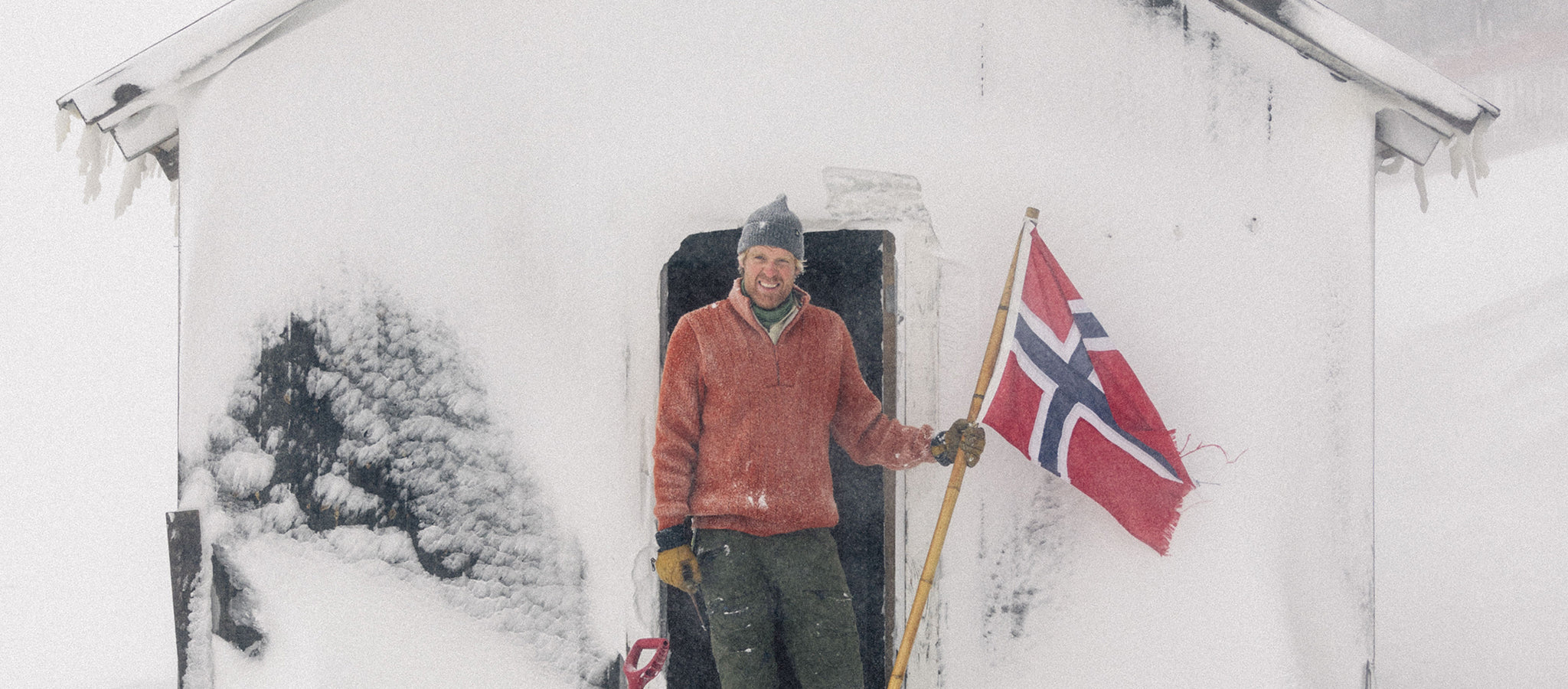 Mann ved hytta på snøfjell, med norsk flagg og Janus Wool Finse genser i farge rød