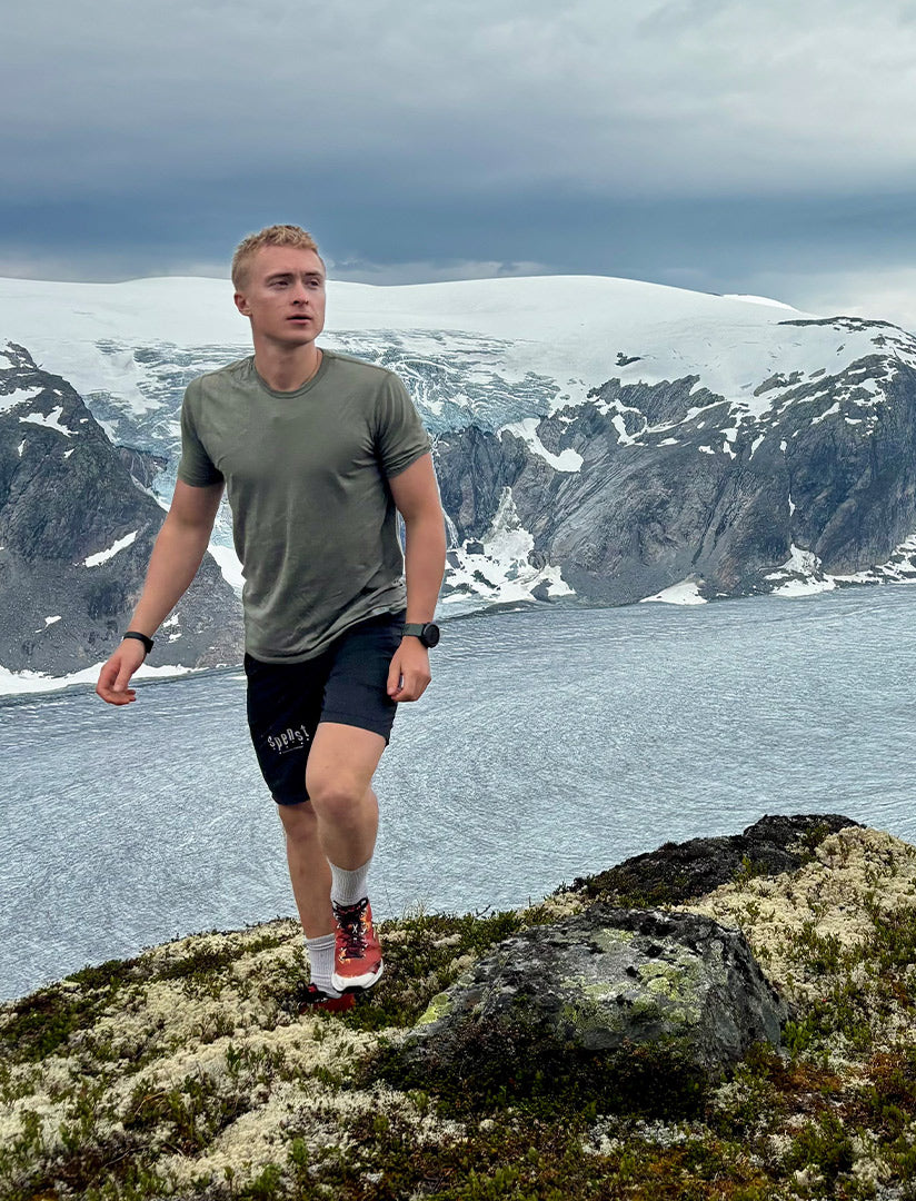 Person standing on a rocky outcrop with a scenic view of mountains and water in the background, wearing Janus merino t-shirt.