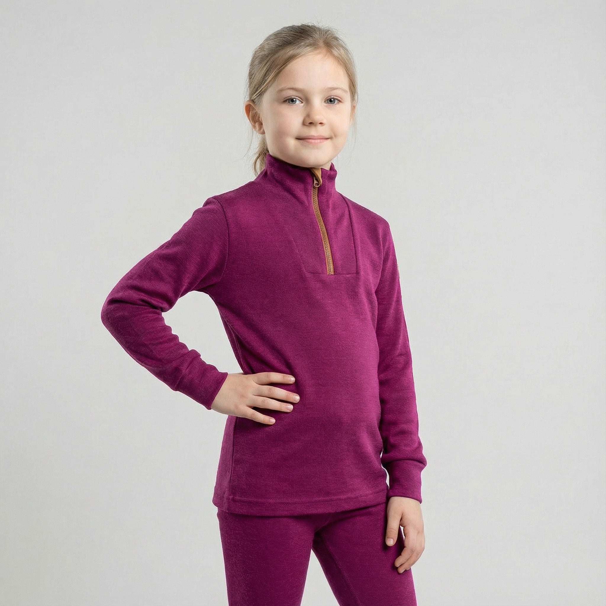 A young girl with light brown hair, wearing a magenta Janus Wool Polo with zip merino - Janus Go 250 Kids and matching pants, stands smiling confidently with one hand on her hip against a plain light gray background.