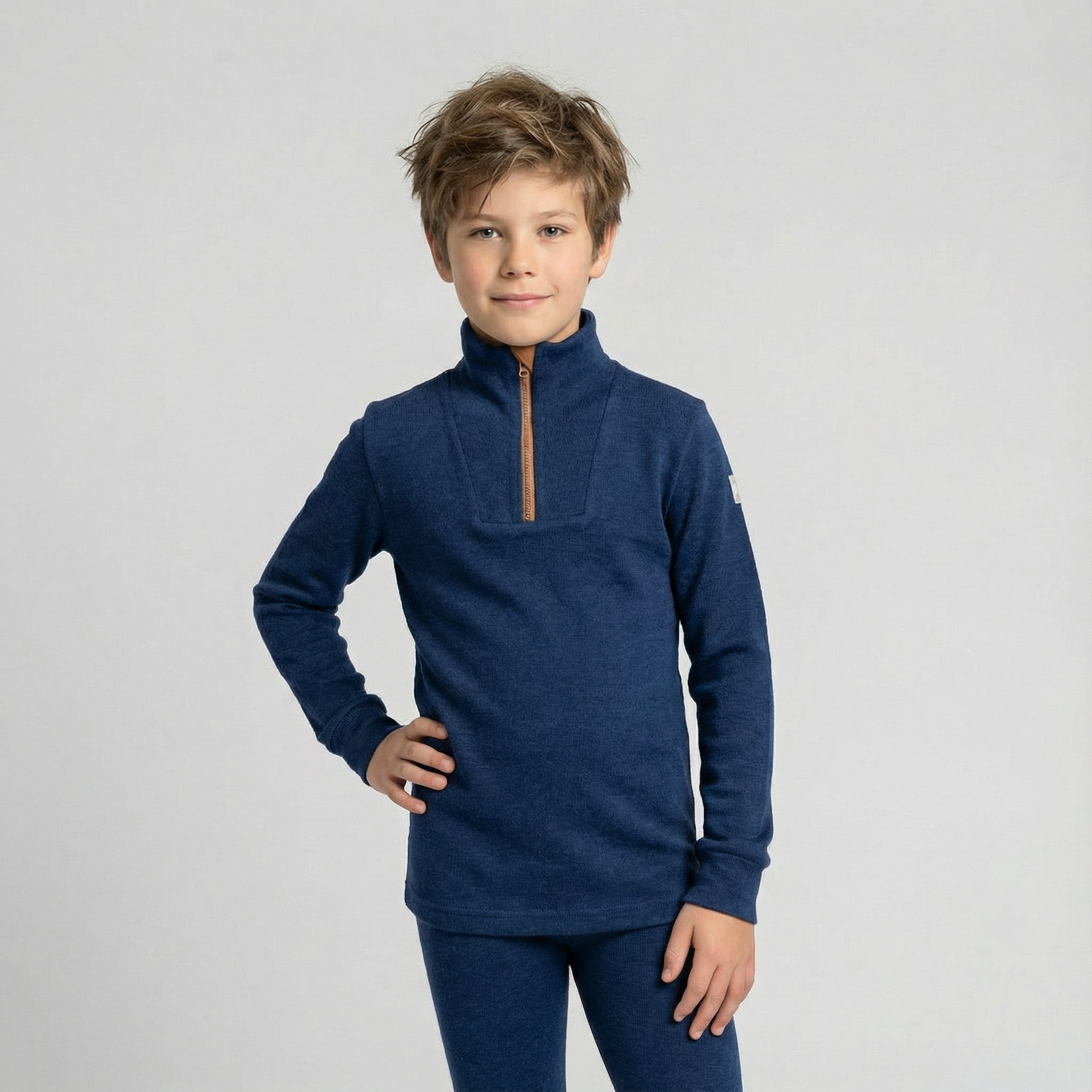 A young boy with light brown hair poses against a gray background, wearing the Janus Wool Polo with zip merino - Janus Go 250 Kids sweater in blue and matching pants. He stands smiling slightly, one hand on his hip.