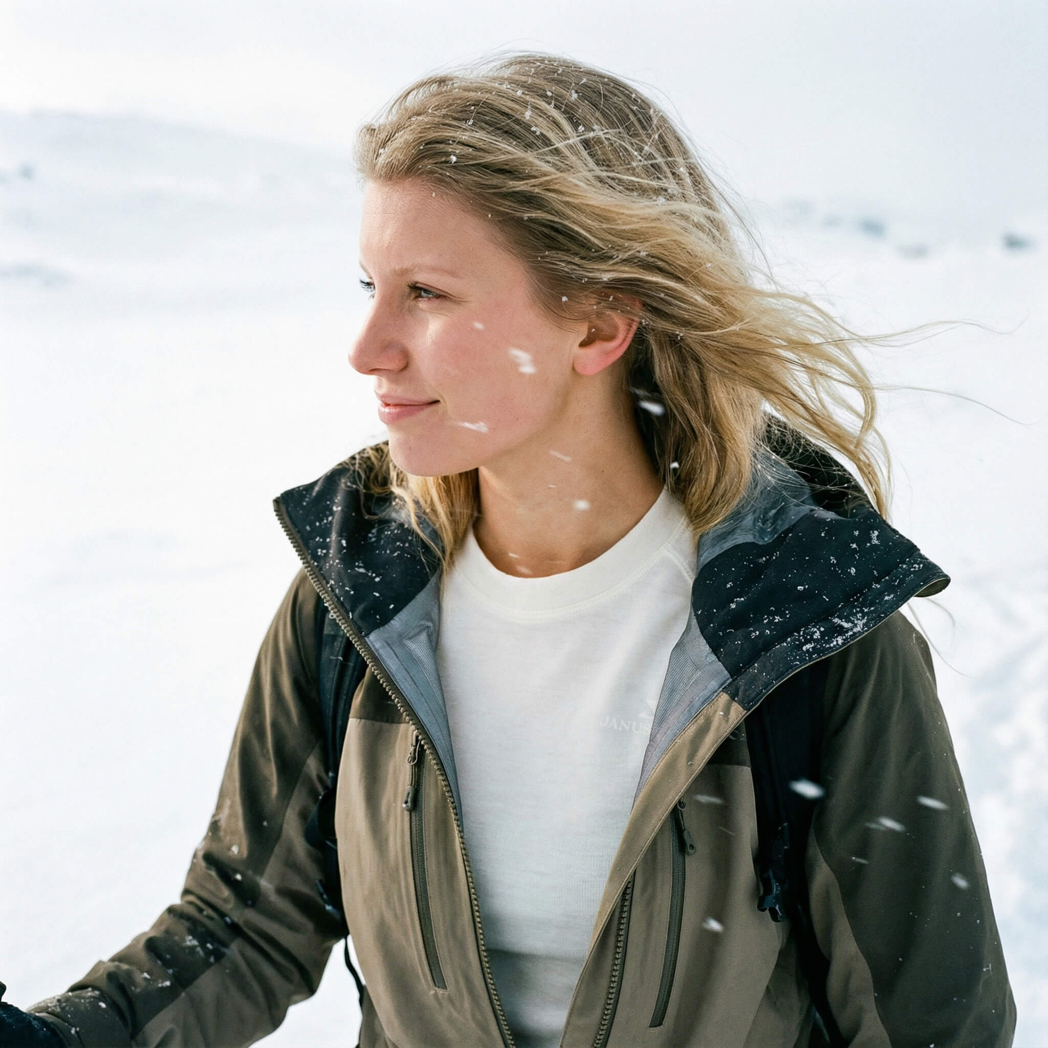 A woman with blonde hair stands outdoors in a snowy landscape, wearing a green merino wool jacket over a white shirt. Snowflakes fall around her as she looks to the side, her hair windblown.