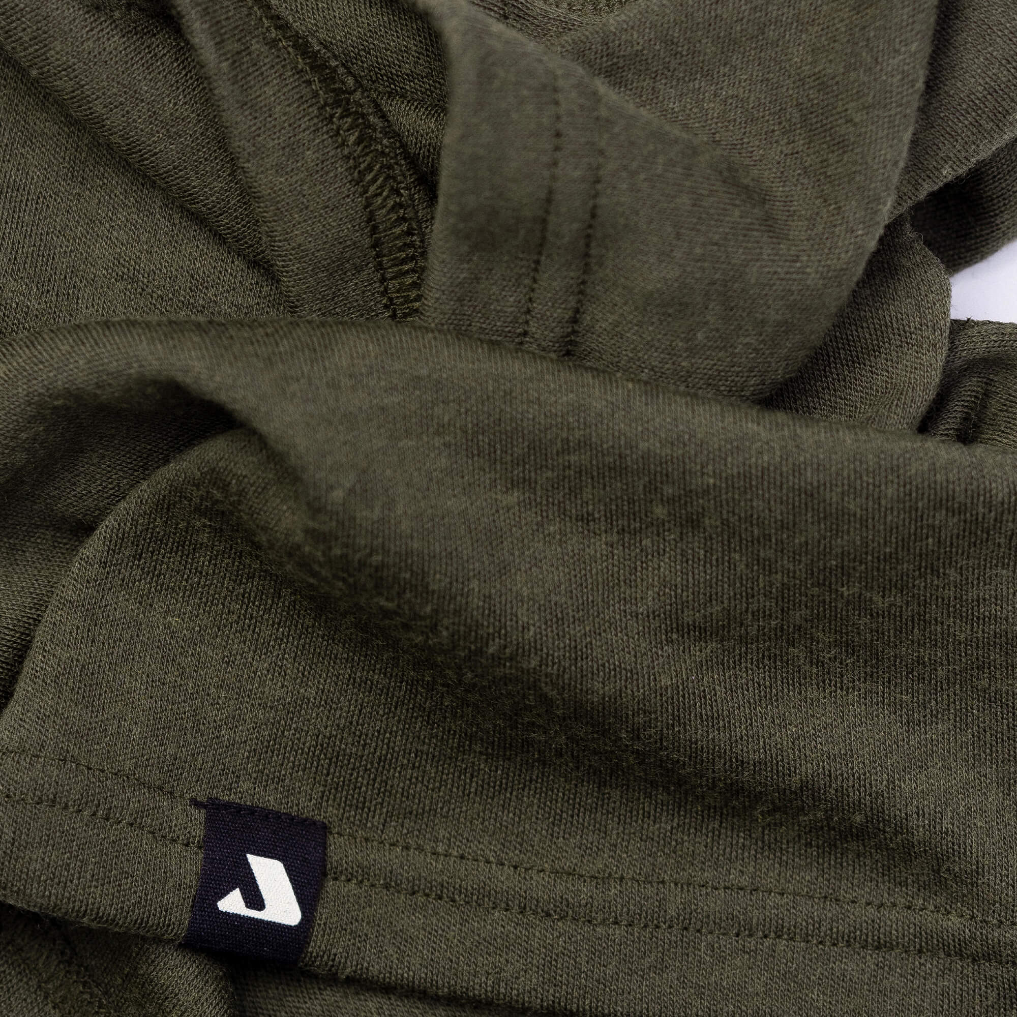 A close-up of olive green fabric with visible stitching and a small black tag featuring a white stylized J near the hem, showcasing the breathable and warm Neck Warmer – Classic Merino 215.