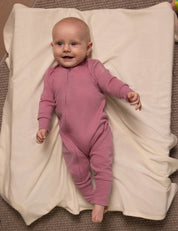 A smiling baby wearing the Pyjamas – Merino Baby Light lies on a white blanket, looking up. The baby has short light hair and is comfortably dressed in breathable merino sleepwear, resting on a textured beige surface.
