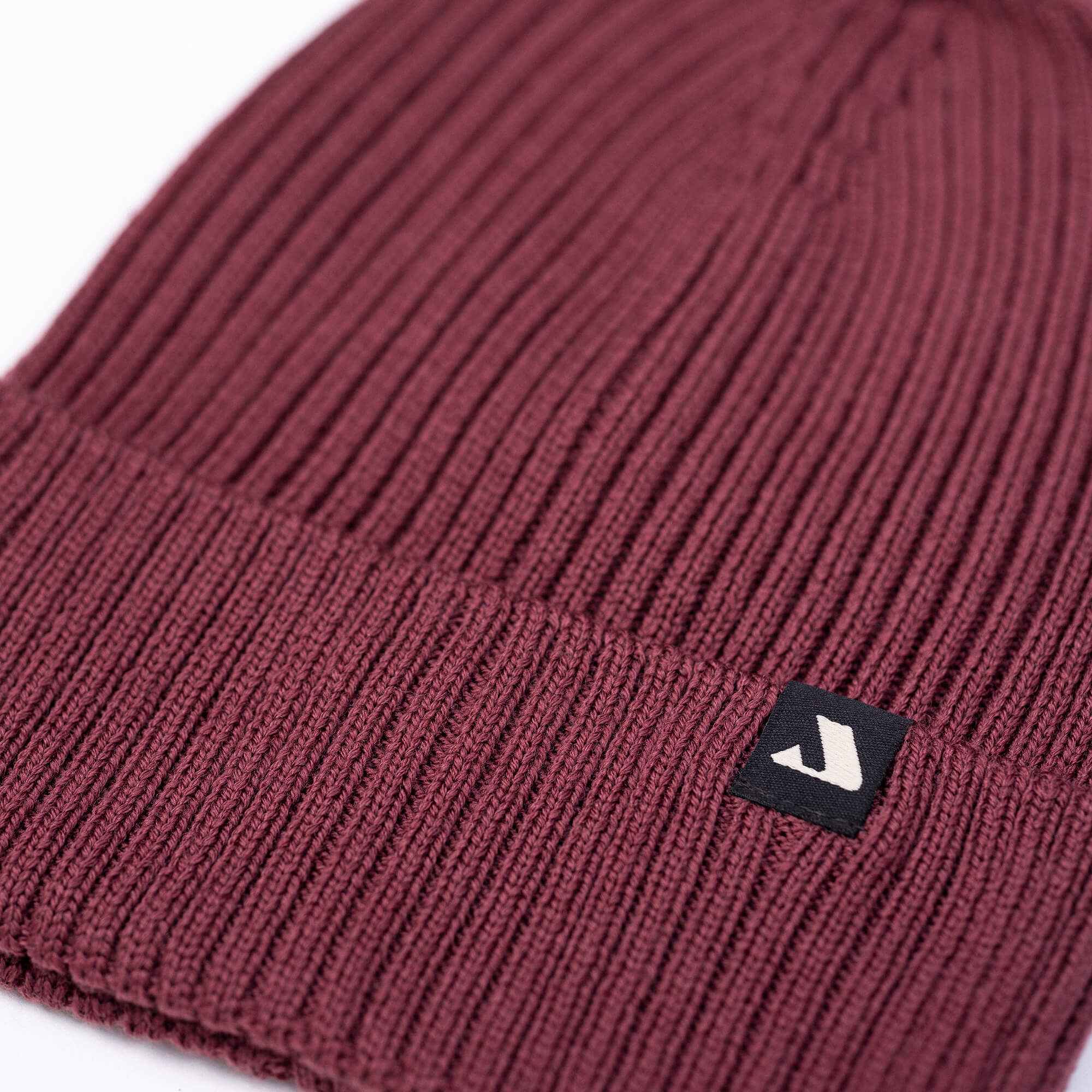 A close-up of the Beanie – Classic in maroon ribbed merino wool with a folded brim and a black tag featuring a white arrow logo, displayed on a white background to emphasize its warmth and breathability.