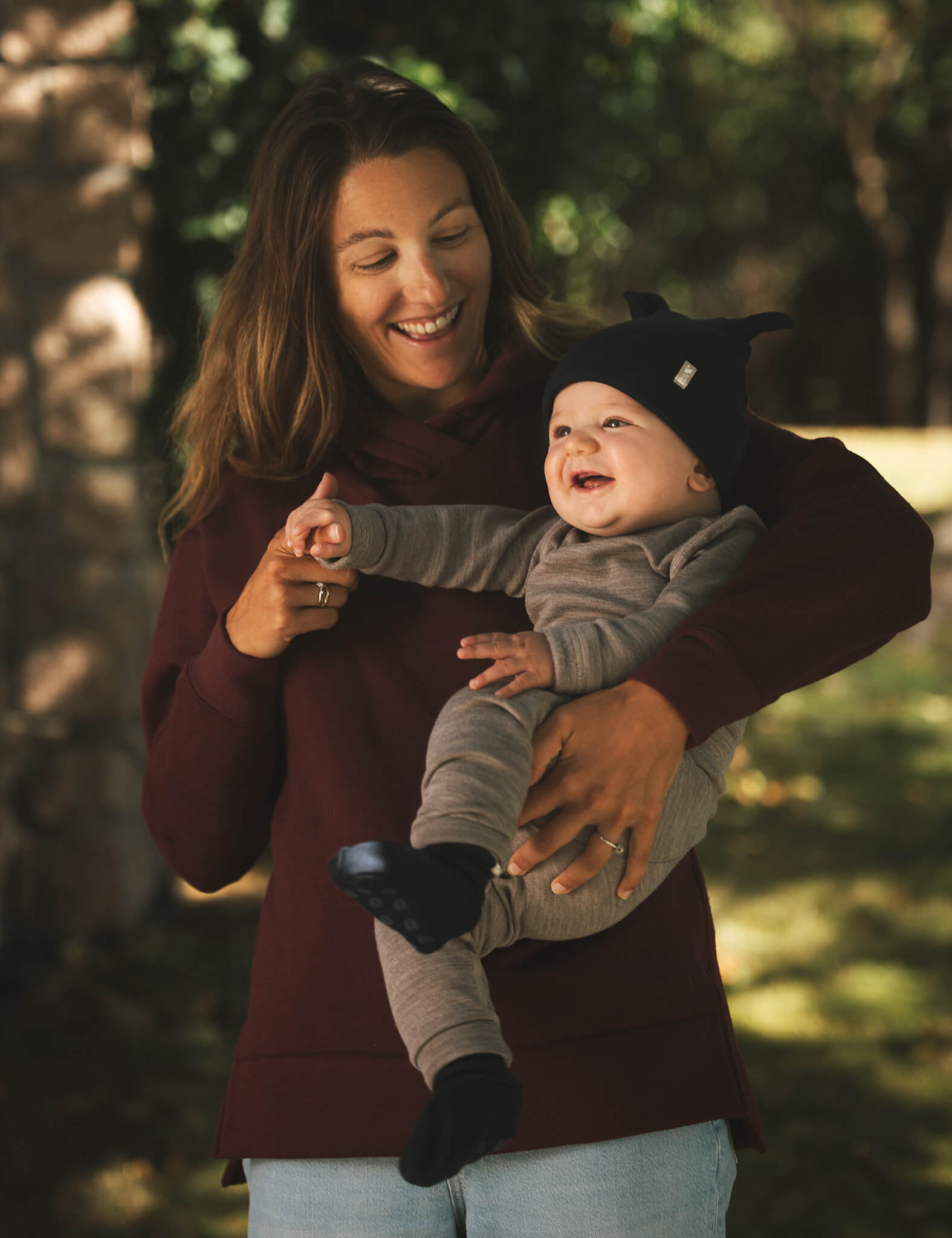 A smiling woman in a maroon hoodie holds a laughing baby dressed in the Merino Baby AirWool Onesie, featuring fold-over cuffs and paired with a black hat. They stand outdoors, surrounded by trees and sunlight.