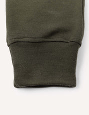 Close-up of an olive green fabric cuff, likely the sleeve or pant leg cuff of a garment such as the Sweatpants – Merino Kids AirWool, shown against a plain light background.