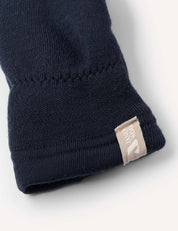 Close-up of the cuff of a navy blue Merino Kids AirWool mitten, featuring a small beige tag with white text. The background is a plain, light color.