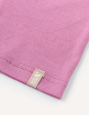 A close-up of the corner of the pink Merino Kids Light Neckwarmer, made from merino wool, features a beige tag with white text and a small design sewn into the seam. The background is white.