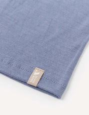 A close-up of the corner of a light blue Neckwarmer – Merino Kids Light, made from merino wool, with a beige tag.