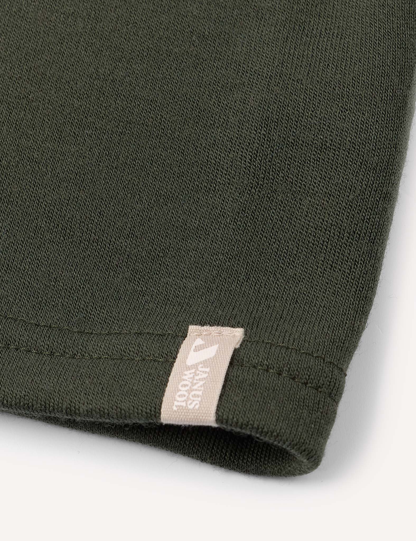 Close-up of the hem of a dark green fabric, likely a garment such as the Neckwarmer – Merino Kids AirWool, featuring a small beige tag with partially visible text and logo near the seam.