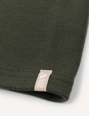 Close-up of the hem of a dark green fabric, likely a garment such as the Neckwarmer – Merino Kids AirWool, featuring a small beige tag with partially visible text and logo near the seam.
