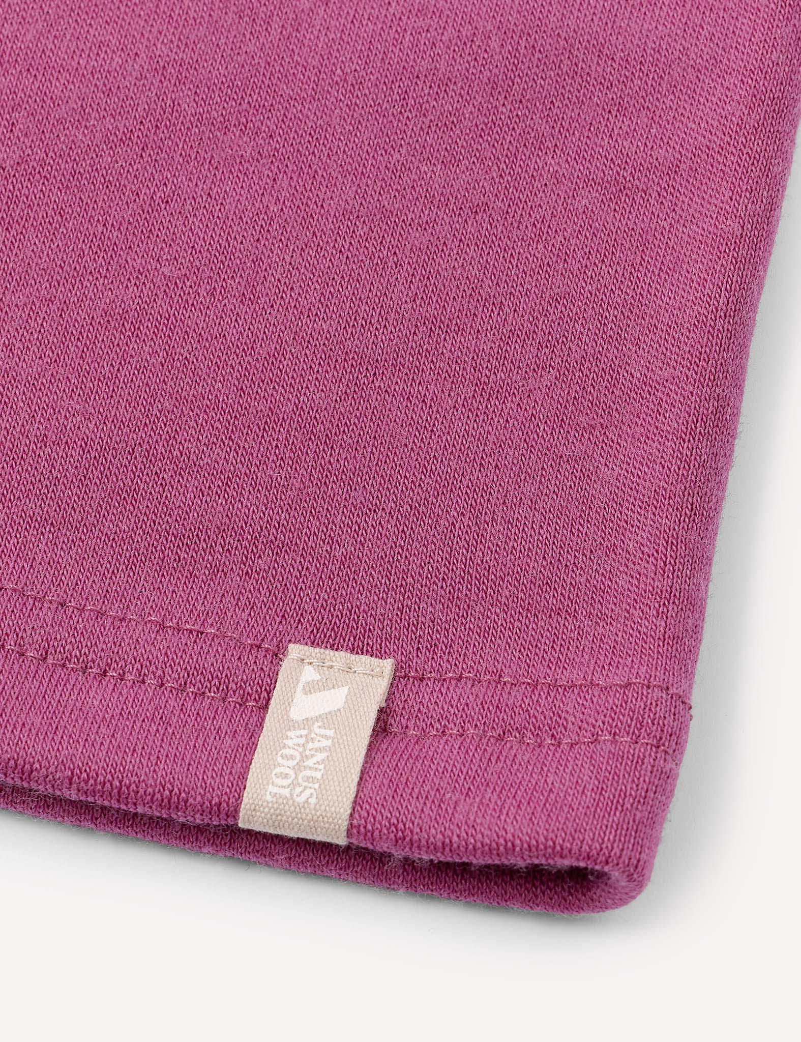 Close-up of a mauve-colored fabric, likely a T-shirt, featuring a beige tag with white text and a small graphic logo sewn into the hem. Set against a light neutral background. Perfect for pairing with the Neckwarmer – Merino Kids AirWool for added comfort and style.