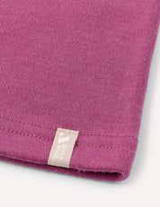 Close-up of a mauve-colored fabric, likely a T-shirt, featuring a beige tag with white text and a small graphic logo sewn into the hem. Set against a light neutral background. Perfect for pairing with the Neckwarmer – Merino Kids AirWool for added comfort and style.