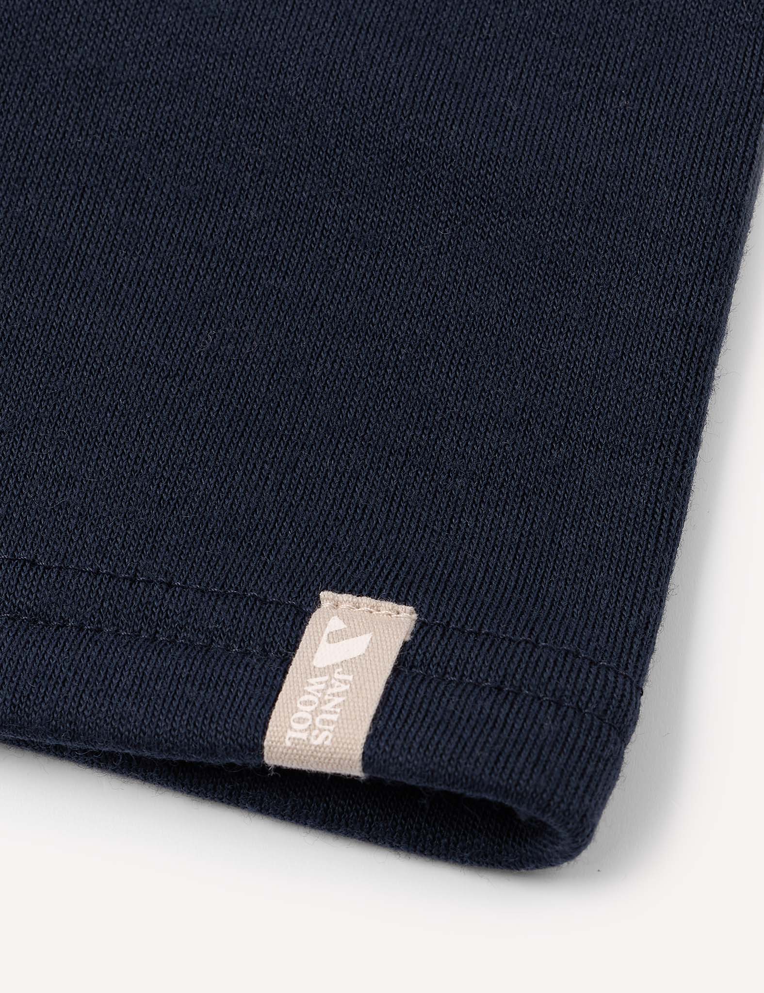 Close-up of the hem of a dark blue knitted fabric garment, likely the Neckwarmer – Merino Kids AirWool, featuring a small beige tag with white text and a logo. The background is plain and light-colored.