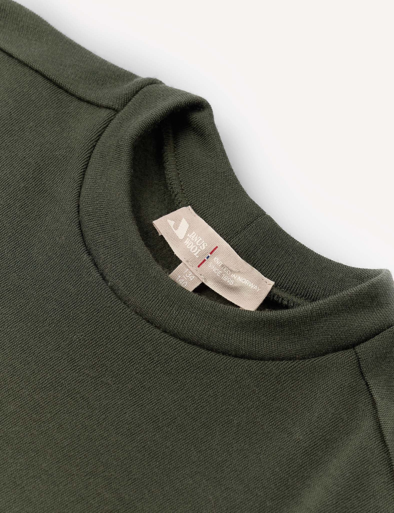 Close-up of an olive green Pullover – Merino Kids AirWool, highlighting the crewneck collar and a beige size tag with red and white accents, set against a white background. This flexible sweater, crafted from AirWool 260 merino wool, exemplifies quality construction and comfort.