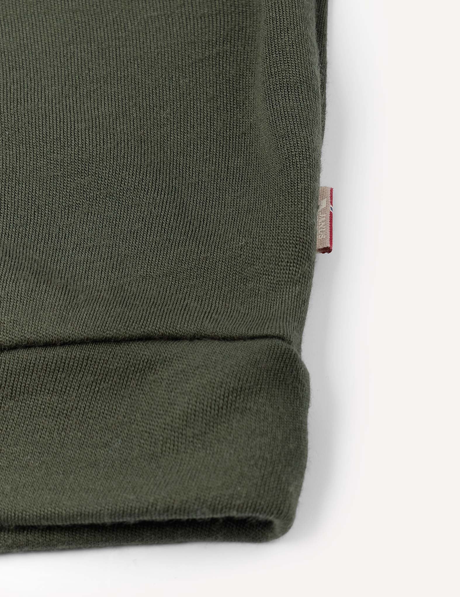 Close-up of the cuff of the Pullover – Merino Kids AirWool in olive green merino wool, featuring a small, partially visible red and white fabric tag sewn into the seam. The background is off-white.