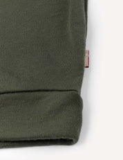 Close-up of the cuff of the Pullover – Merino Kids AirWool in olive green merino wool, featuring a small, partially visible red and white fabric tag sewn into the seam. The background is off-white.
