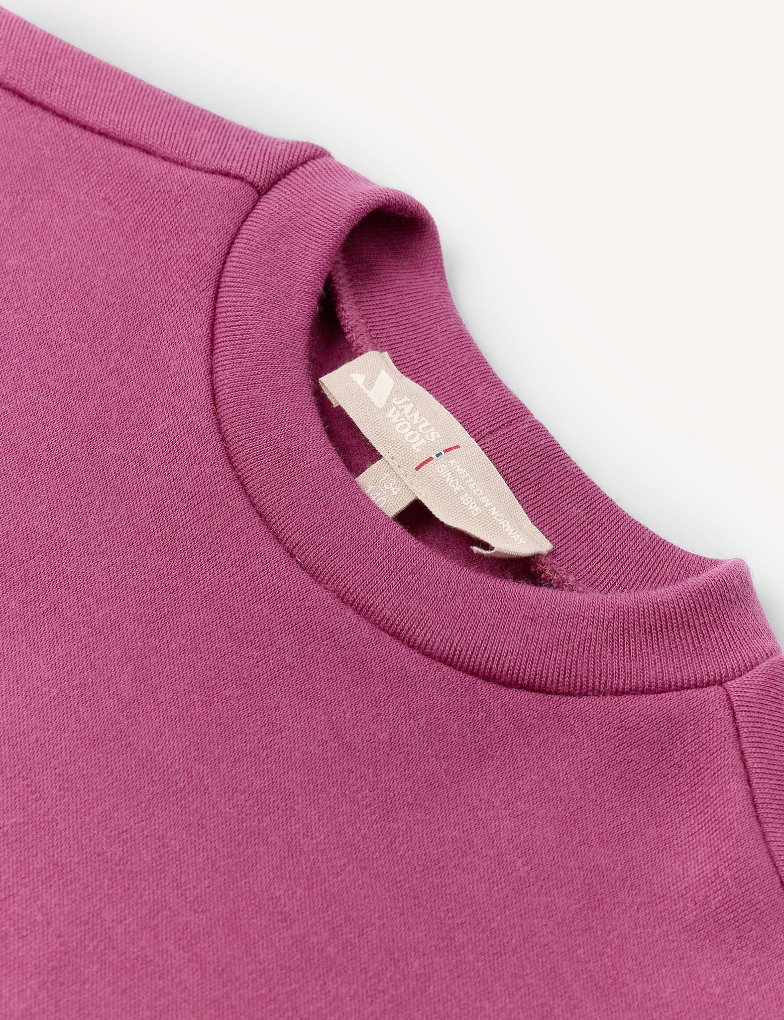 A close-up of the Pullover – Merino Kids AirWool in magenta, highlighting the crewneck neckline and a beige tag featuring text and a small red stitch, set against a light background.