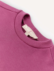 A close-up of the Pullover – Merino Kids AirWool in magenta, highlighting the crewneck neckline and a beige tag featuring text and a small red stitch, set against a light background.