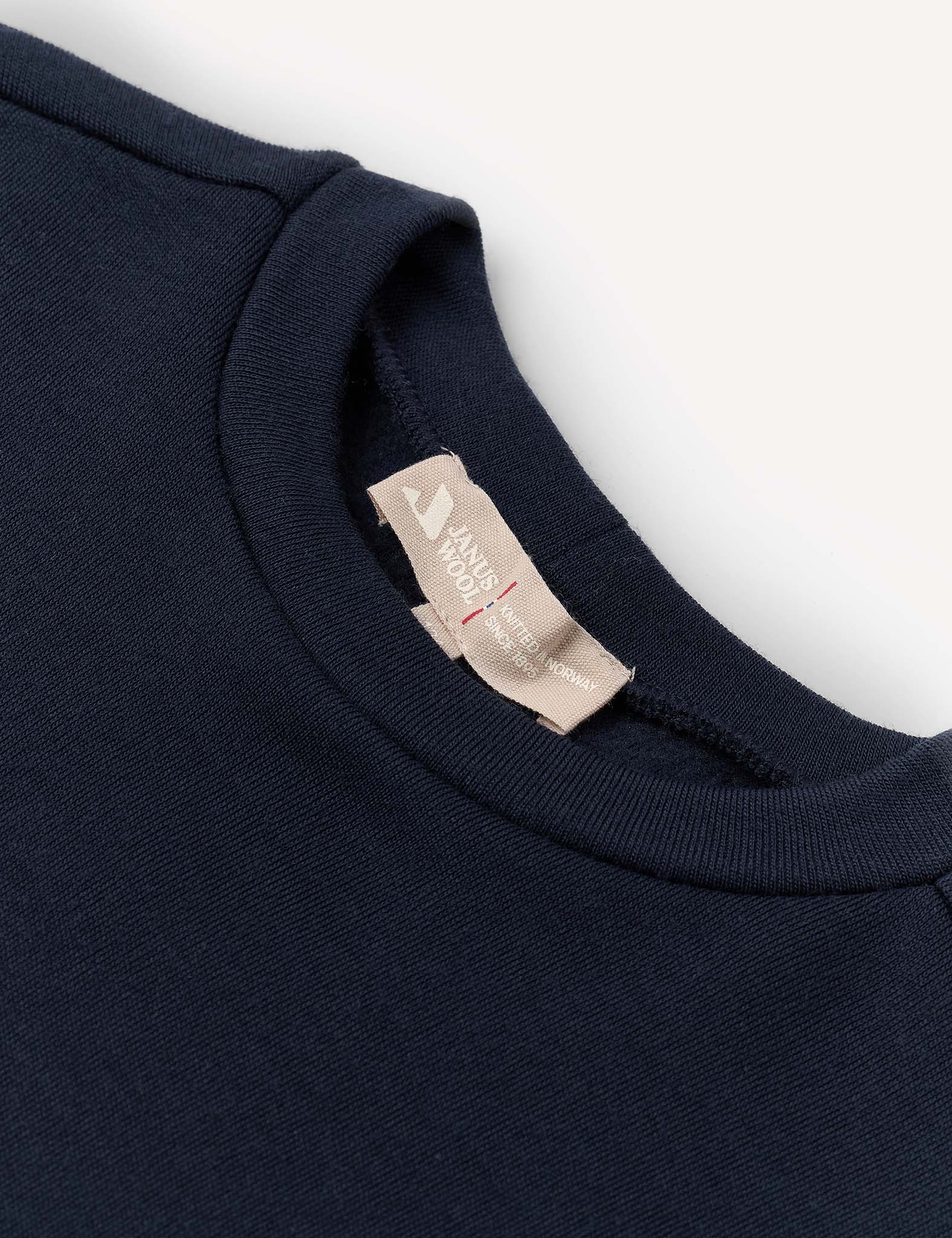 A close-up of the neckline and collar of a navy blue crewneck shirt, featuring a beige fabric tag with text and a red stitched line, set against a white background. This Pullover – Merino Kids AirWool sweater highlights the fine details and craftsmanship of the collection.