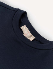 A close-up of the neckline and collar of a navy blue crewneck shirt, featuring a beige fabric tag with text and a red stitched line, set against a white background. This Pullover – Merino Kids AirWool sweater highlights the fine details and craftsmanship of the collection.
