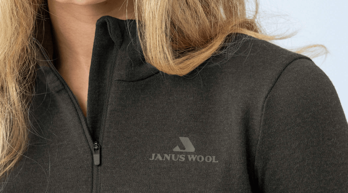 A close-up of a person wearing a dark zip-up merino wool jacket with the WOOL logo on the chest. The person's long blonde hair partially covers their face.
