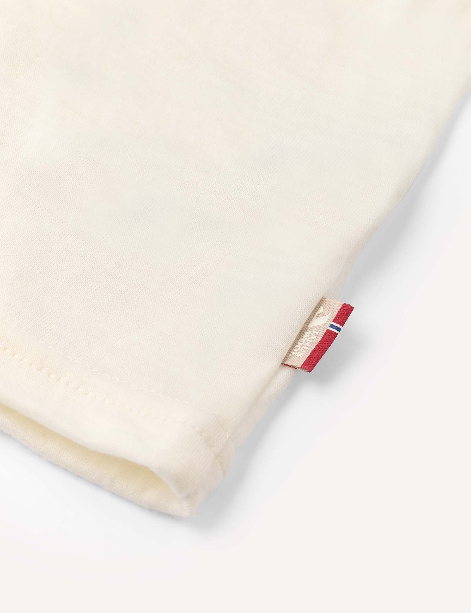 Close-up of the hem of the cream-colored “T-Shirt – Merino Kids Light,” featuring a small beige and red tag sewn into the seam on a light background. Made from merino wool, this fabric is celebrated for its natural temperature regulation and all-day comfort.