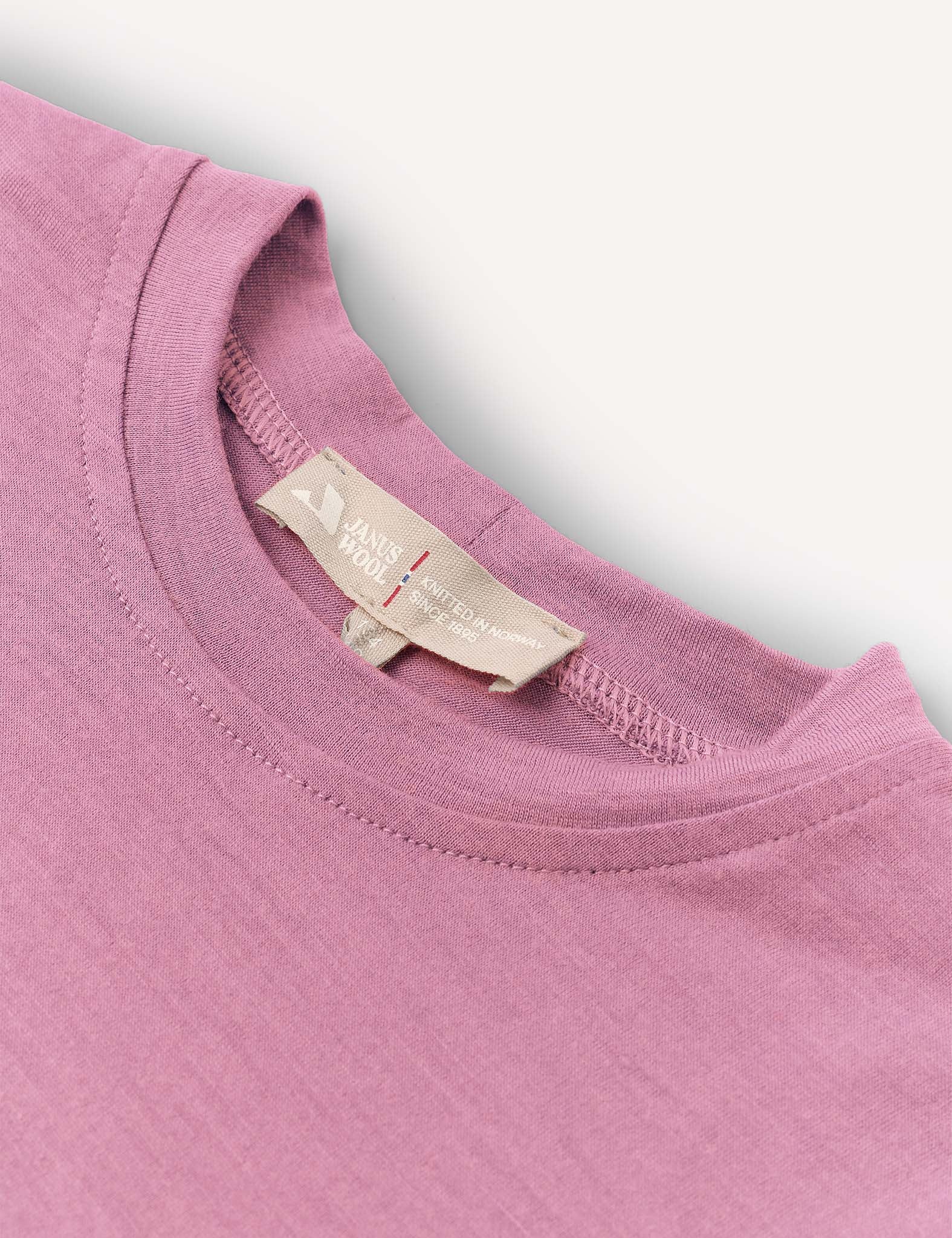 A close-up of the neckline and label of the T-Shirt – Merino Kids Light in mauve merino wool. Celebrated for its natural temperature-regulating properties and all-day comfort, the shirt is shown laid flat on a white surface.