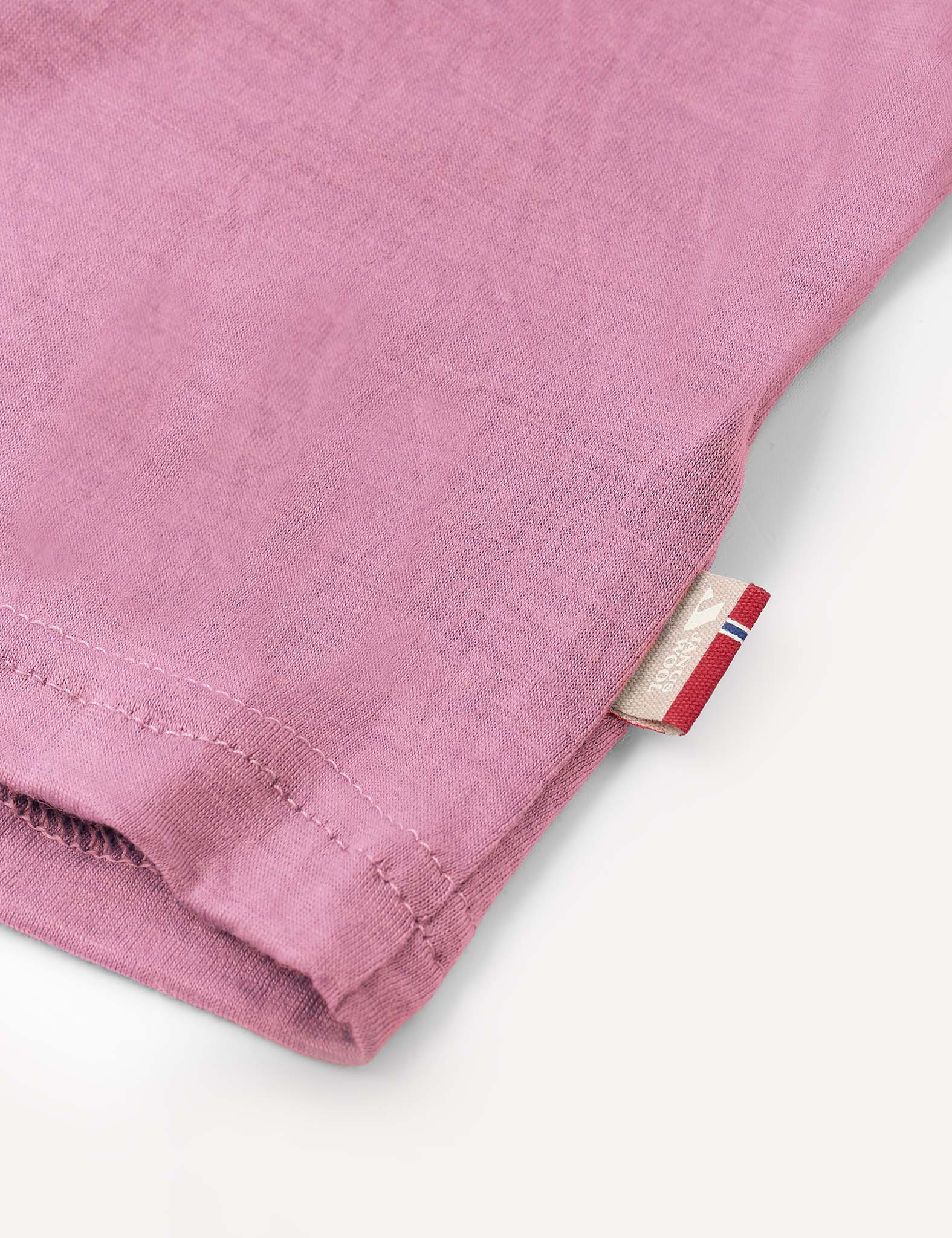 A close-up of the T-Shirt – Merino Kids Light in pink, featuring a hemmed edge and a small tag with red, white, and blue stripes and partial text. Crafted from merino wool for all-day comfort and natural temperature regulation.