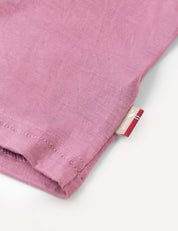 A close-up of the T-Shirt – Merino Kids Light in pink, featuring a hemmed edge and a small tag with red, white, and blue stripes and partial text. Crafted from merino wool for all-day comfort and natural temperature regulation.