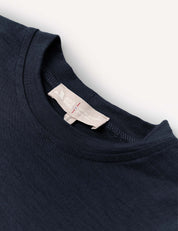 Close-up of the T-Shirt – Merino Kids Light in navy blue merino wool, featuring a beige collar tag. Renowned for its natural temperature regulation and all-day comfort, the shirt is laid flat on a white surface.