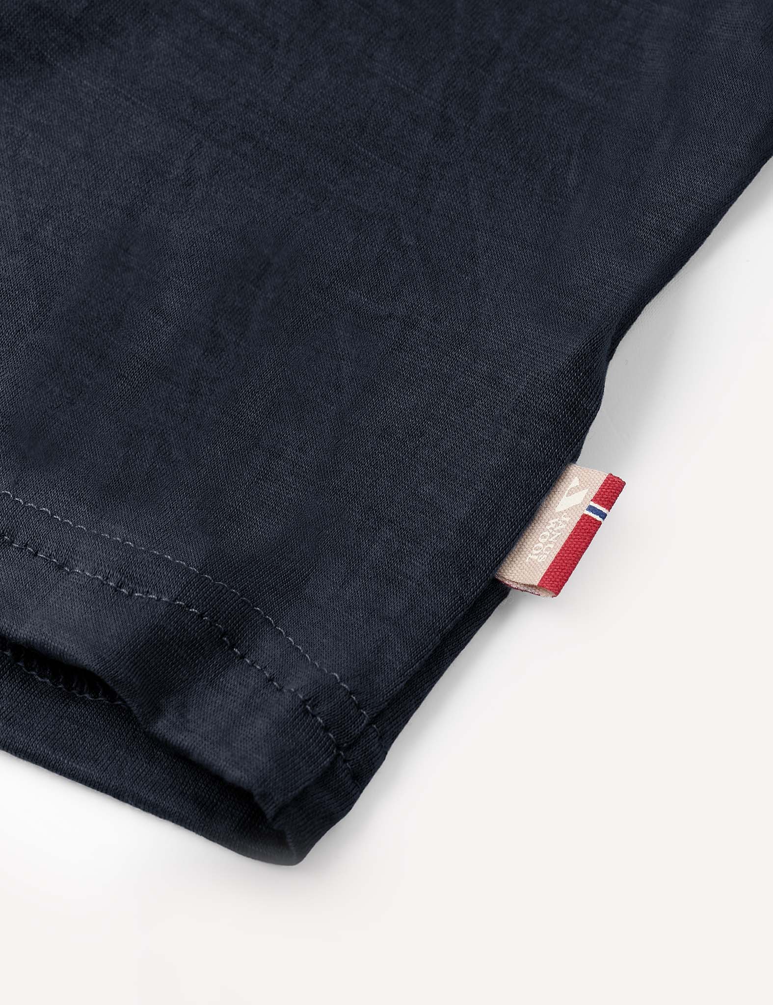 Close-up of the hem of the T-Shirt – Merino Kids Light in dark blue merino wool, featuring a small beige tag with red, white, and blue details stitched into the side seam. The fabric delivers natural temperature regulation and all-day comfort.