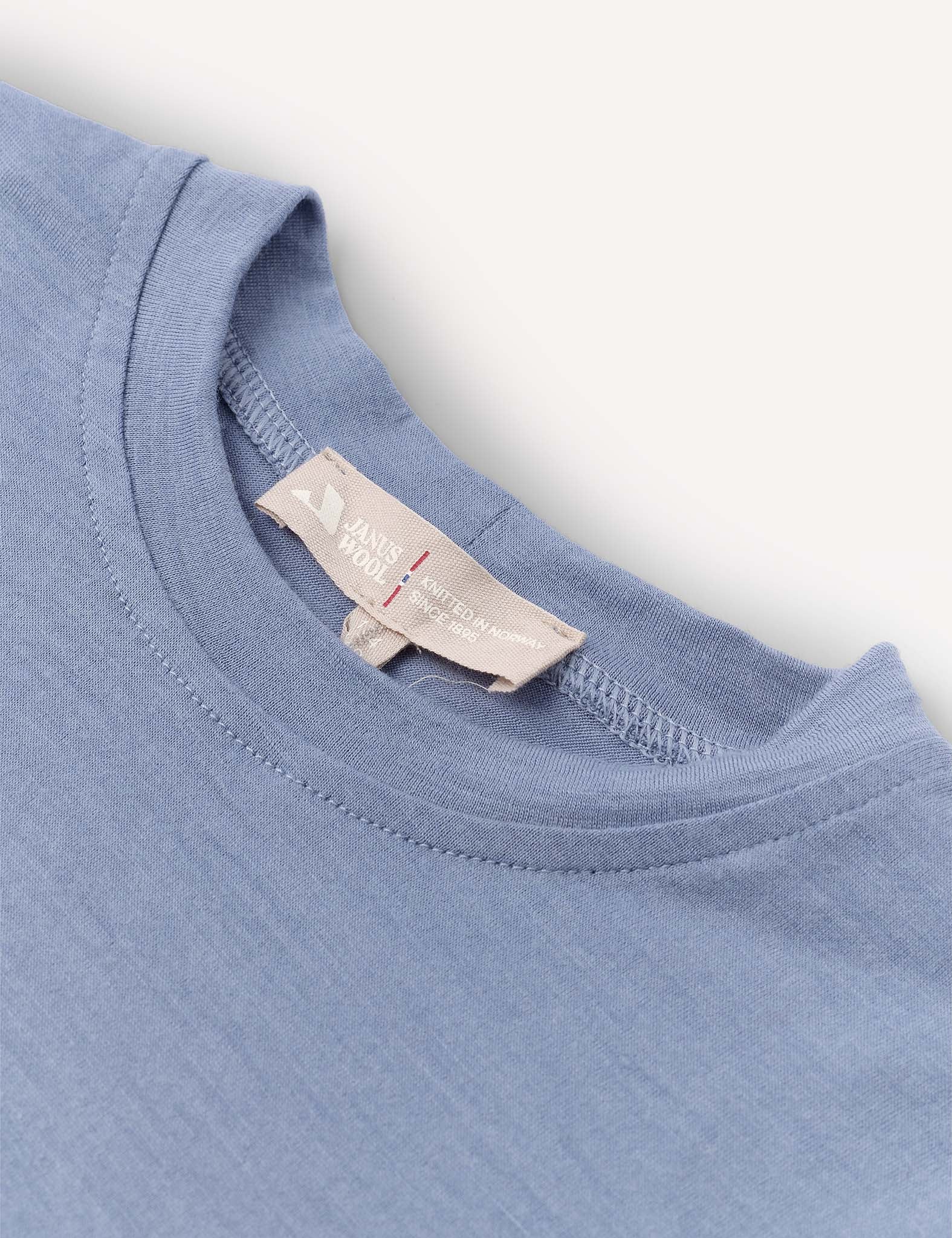 Close-up of a T-Shirt – Merino Kids Light in light blue, highlighting the collar and a beige tag featuring the brand name and care instructions. The detailed view of the merino wool fabric texture and precise stitching emphasizes its natural temperature-regulating properties for all-day comfort.