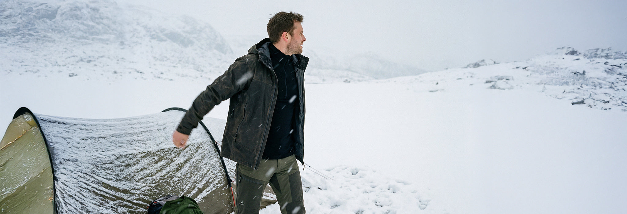 A man stands outside a tent in a snowy landscape, dressed in a dark merino wool jacket and pants. Snow blankets the ground and tent, with mountains visible in the background beneath an overcast sky.