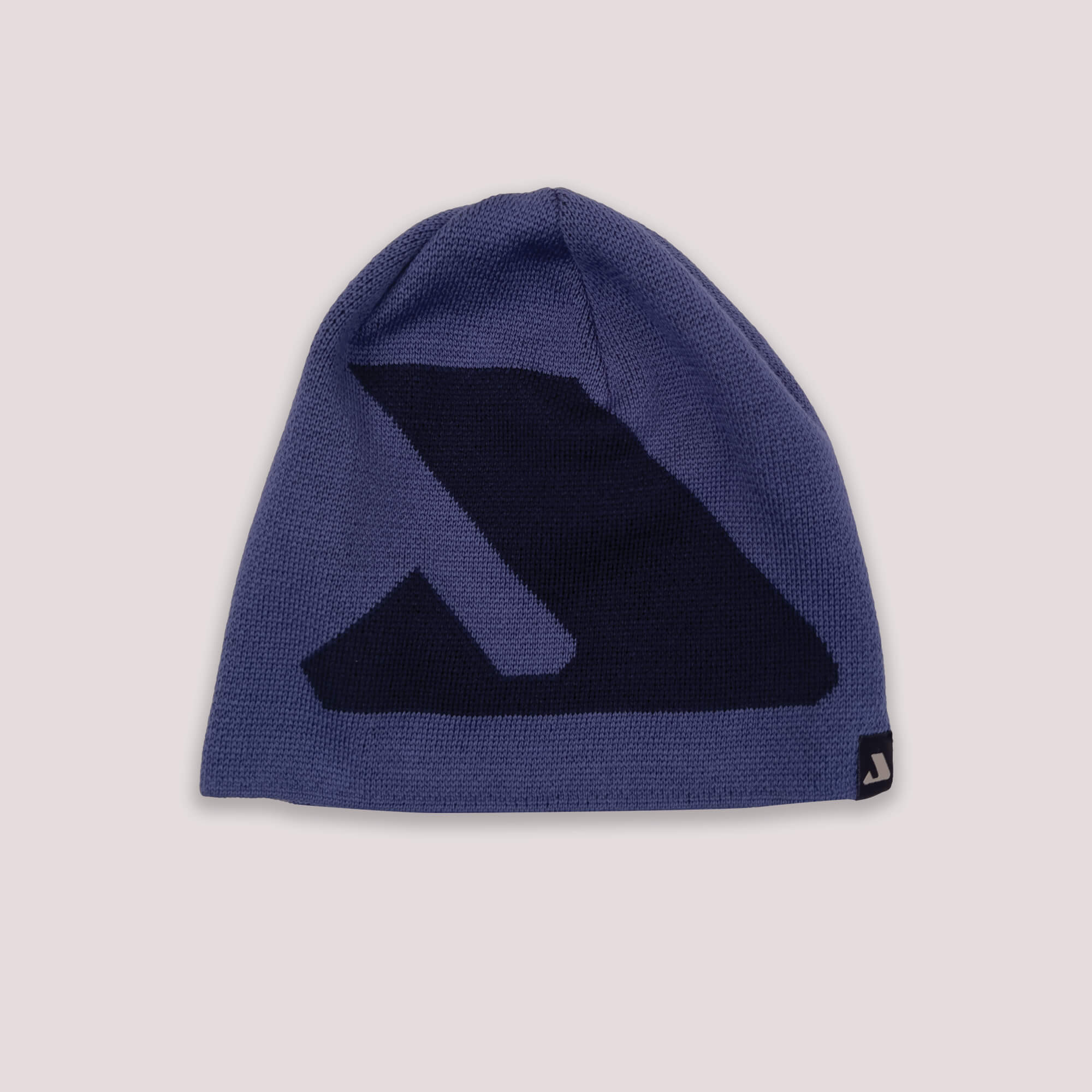 The Beanie – Logo in Skipper Blue features a bold black geometric design on the front and a small black tag with white logo at the edge, providing warmth and breathability. Shown against a light pink background.